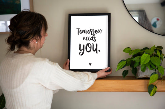 Person holding a framed quote 'Tomorrow needs you' on a shelf with a plant in the background