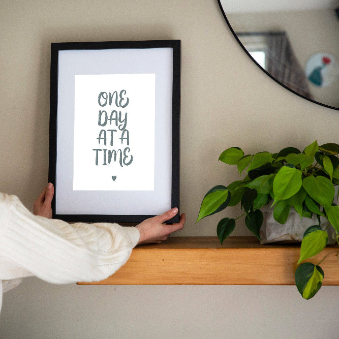 One Day at a Time typographic print in sage on white framed in black held by brunette woman with plant beside and mirror on wall, by Paper Heart Design NZ