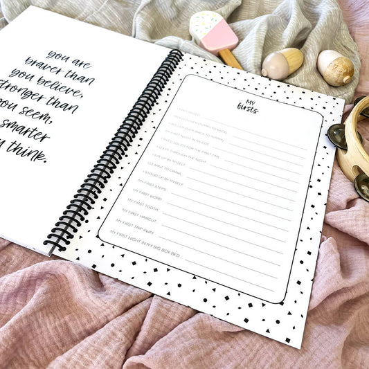 Personalised Hey Baby milestone book open to quote and My Firsts pages flat lay on pink and grey background with wooden baby toys, by Paper Heart Design NZ