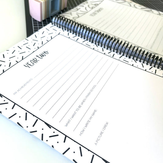 Personalised My School Days school record book close up of blank year two page with year three page behind, sitting on black grid mat and white surface, by Paper Heart Design NZ