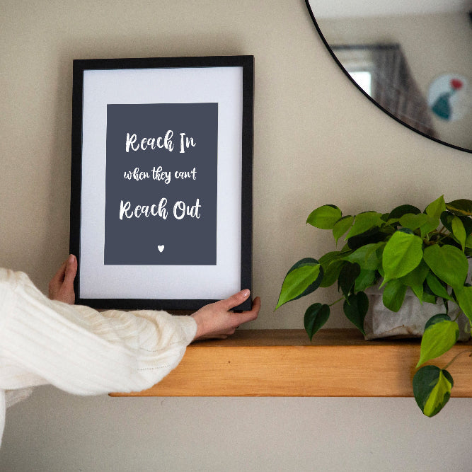Reach In When They Can't Reach Out print in dusk framed in black with white matt held by brunette woman with shelf, mirror and plant, by Paper Heart Design NZ