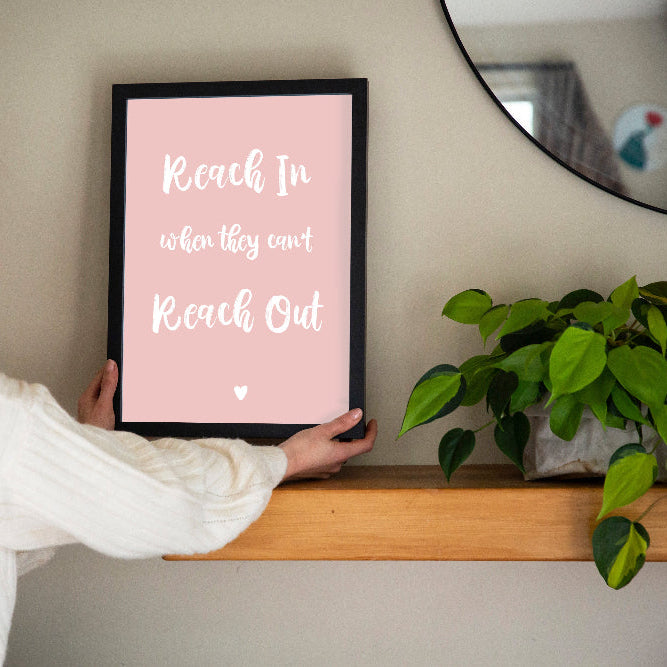 Reach In When They Can't Reach Out print in marshmallow pink framed in black held by brunette woman with shelf, mirror and plant, by Paper Heart Design NZ