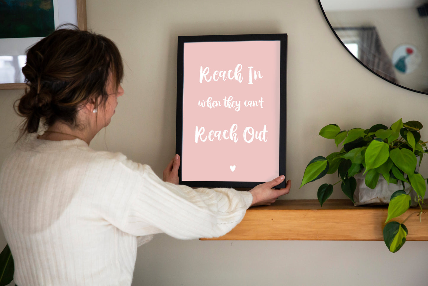 Reach In When They Can't Reach Out print in marshmallow pink framed in black held by brunette woman with shelf, mirror and plant, by Paper Heart Design NZ