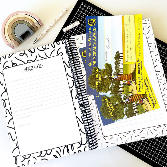 School Record Book open to year one pages showing heavy duty premium pocket with certificates and pen, flat lay on black grid mat and white surface, by Paper Heart Design NZ