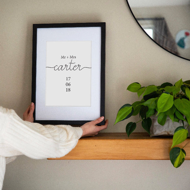 You and Me Wedding Date print in black frame held by brunette woman over shelf with mirror and plant behind, personalised wedding wall art by Paper Heart Design NZ