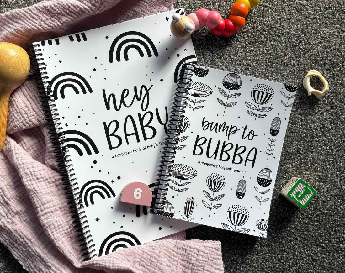 Paper Heart Design Spiral Bound Keepsake Books Hey Baby and Bump to Bubba sitting on a grey and pink textured surface, surrounded by wooden baby toys