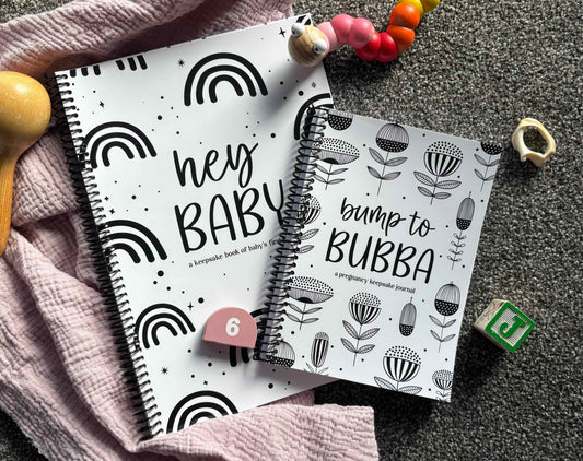 Paper Heart Design Spiral Bound Keepsake Books Hey Baby and Bump to Bubba sitting on a grey and pink textured surface, surrounded by wooden baby toys