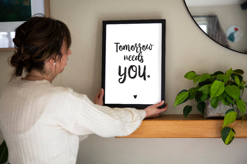 Person holding a framed quote Tomorrow needs you on a shelf with a plant in the background, designed by Paper Heart Design NZ to support suicide prevention and awareness