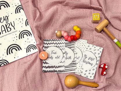  Downloadable baby milestone cards four assorted black and white cards flat lay on pink background with wooden toys and Hey Baby book, by Paper Heart Design