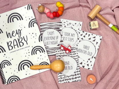 Downloadable baby milestone cards four cards with Hey Baby keepsake book and wooden baby toys flat lay on pink cloth background, by Paper Heart Design