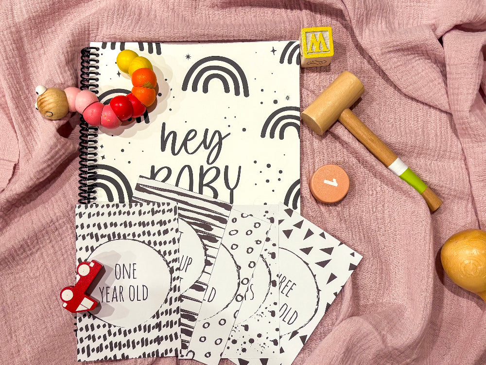 Downloadable baby milestone cards with Hey Baby first year keepsake book flat lay on pink background with baby themed toys, by Paper Heart Design