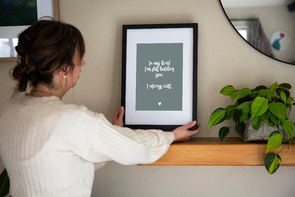 Brunette Lady holding In My Heart memorial print in sage framed on wooden shelf with plant and mirror, memorial keepsake print by Paper Heart Design NZ
