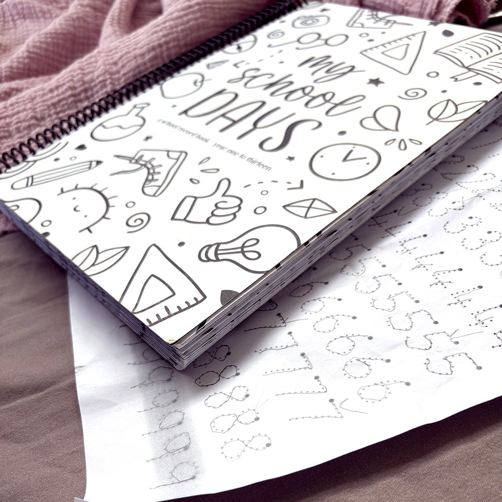 My School Days school record book closed cover with Schoolies design on white background, lying on pink fabric with child's homework sheet visible beneath, showing thickness of keepsake book and quality of paper and binding, by Paper Heart Design NZ