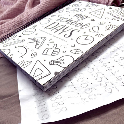 My School Days school record book closed cover with Schoolies design on white background, lying on pink fabric with child's homework sheet visible beneath, showing thickness of keepsake book and quality of paper and binding, by Paper Heart Design NZ