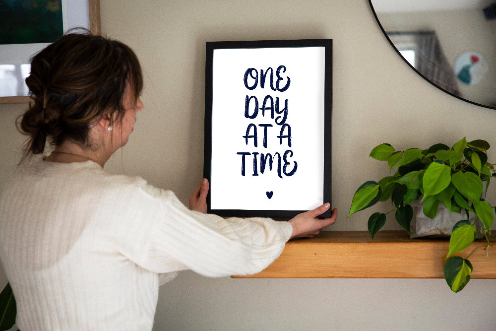 One Day at a Time typographic print in navy framed in black held by brunette woman with plant beside and mirror on wall, by Paper Heart Design NZ
