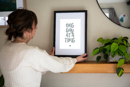 One Day at a Time typographic print in sage on white framed in black held by brunette woman with plant beside and mirror on wall, by Paper Heart Design NZ