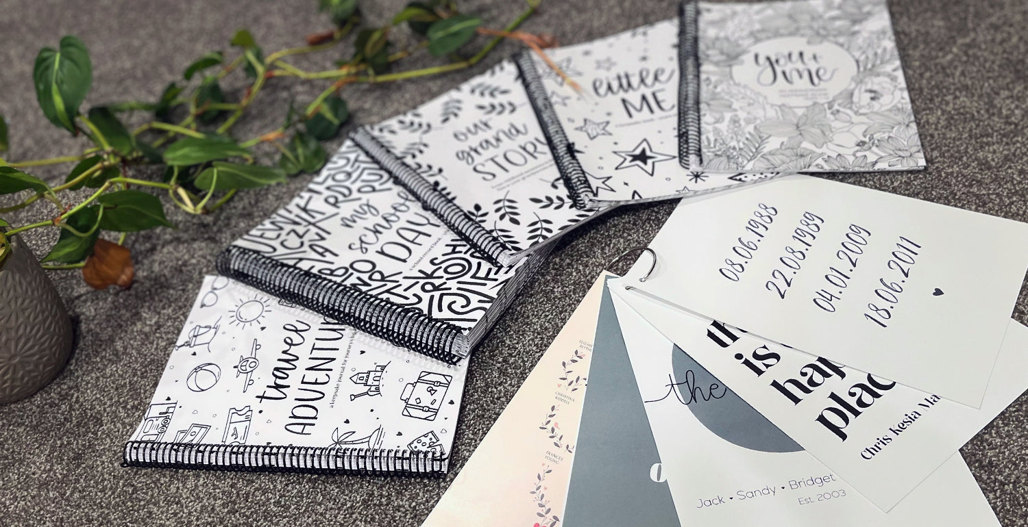 Flat lay of a selection of Paper Heart Design's keepsake books and personalised family prints, including school record books, milestone journals, and family tree prints, styled with greenery