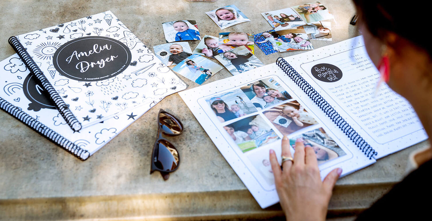 female writing in baby milestone book at concrete table, surrounded by photos and other keepsake journals, made by Paper Heart Design