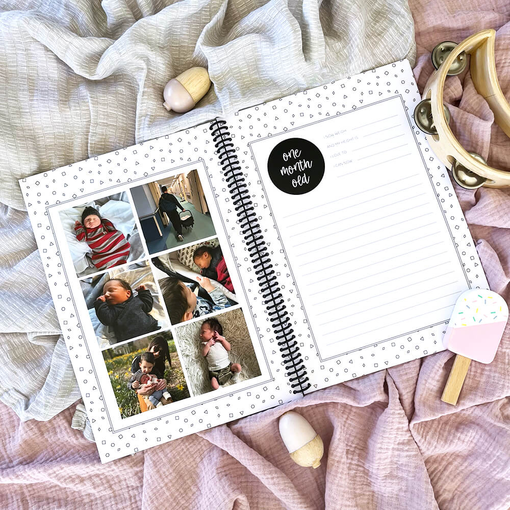 Personalised Hey Baby milestone book open to one month journal page with baby photo pages opposite, flat lay on pink and grey background with wooden toys, by Paper Heart Design NZ