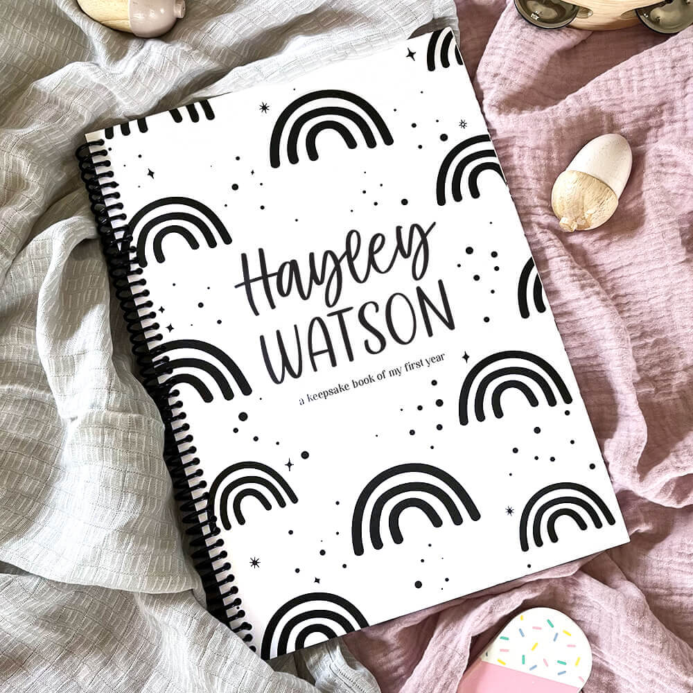 Personalised Hey Baby milestone book with rainbows cover showing child's name on pink and grey textured fabric, custom baby keepsake by Paper Heart Design NZ