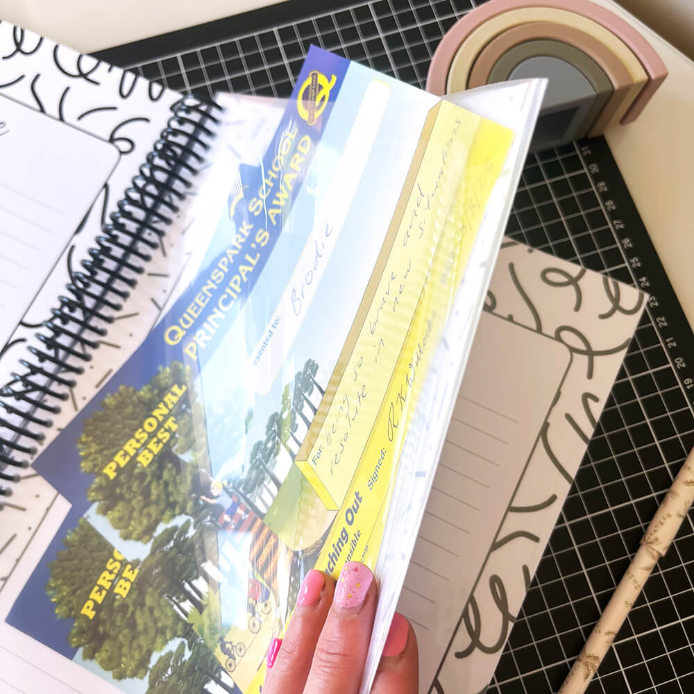 Personalised My School Days school record book close up of year page pocket holding certificates, held open by female hand with pink nails on black grid mat and white surface, by Paper Heart Design NZ