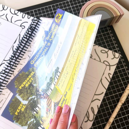 Personalised My School Days school record book close up of year page pocket holding certificates, held open by female hand with pink nails on black grid mat and white surface, by Paper Heart Design NZ