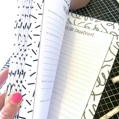 Personalised My School Days school record book open to year thirteen page and empty pocket, held partially open by female hand with pink nails, by Paper Heart Design NZ
