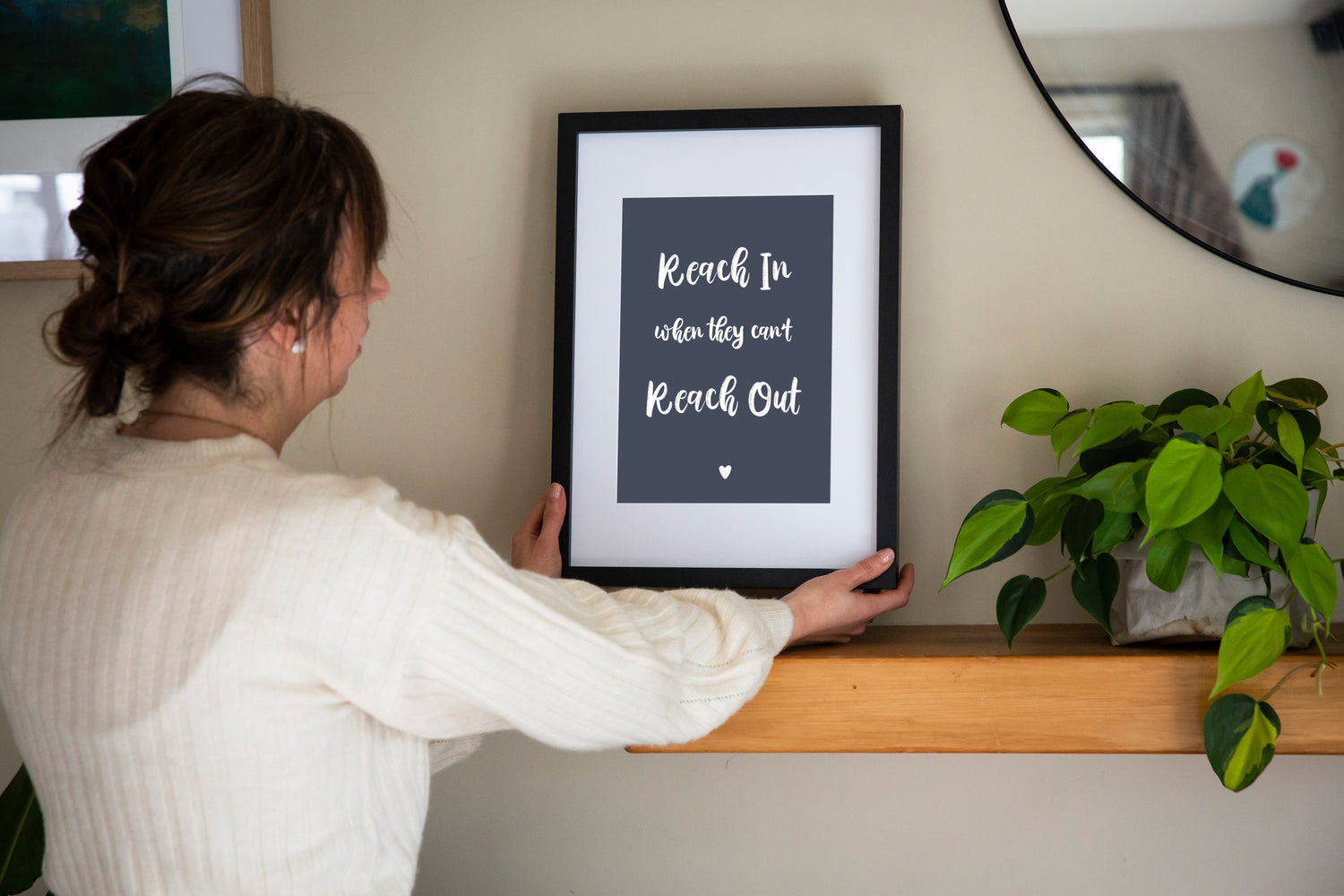 Reach In When They Can't Reach Out print in dusk framed in black with white matt held by brunette woman with shelf, mirror and plant, by Paper Heart Design NZ
