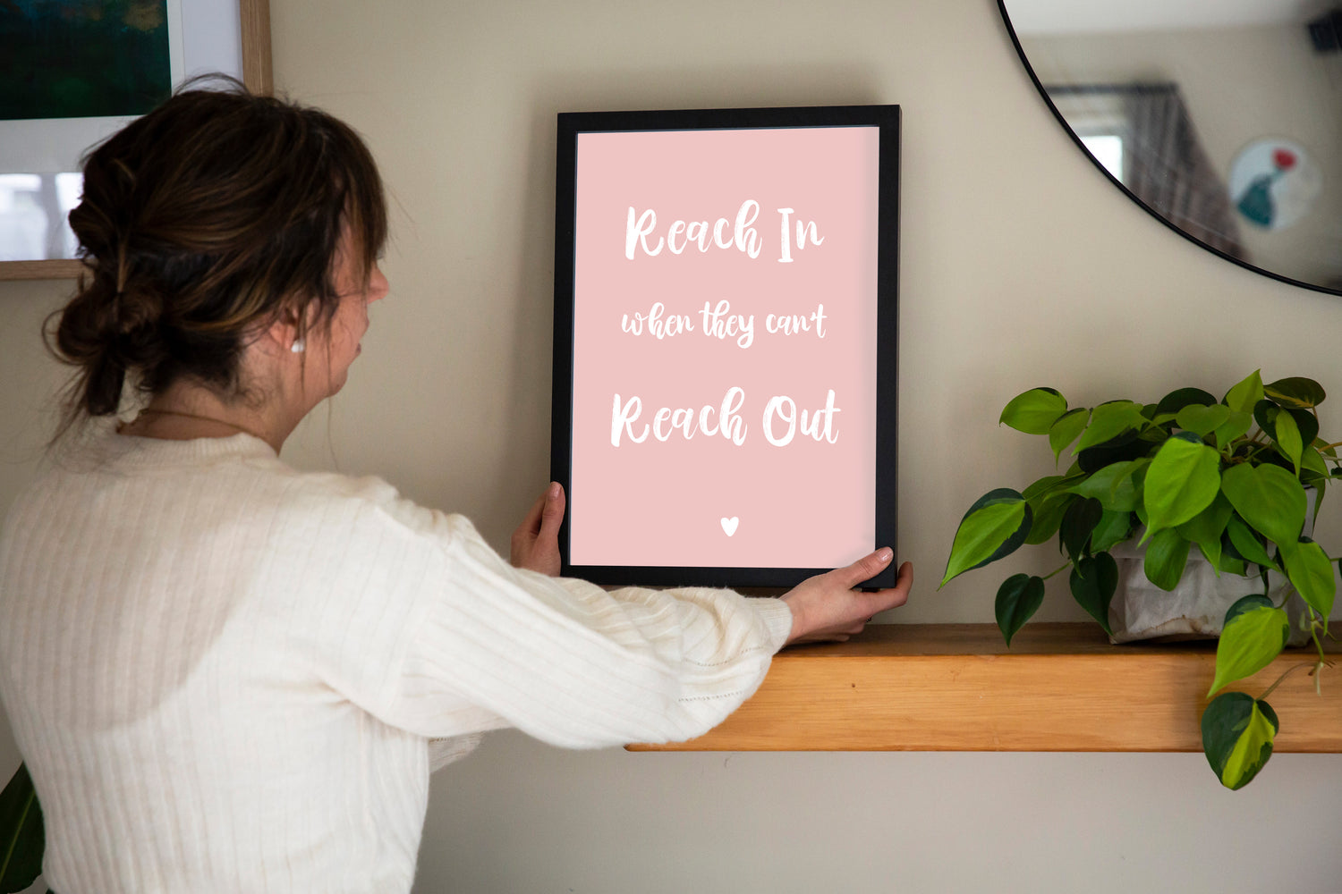 Reach In When They Can't Reach Out print in marshmallow pink framed in black held by brunette woman with shelf, mirror and plant, by Paper Heart Design NZ