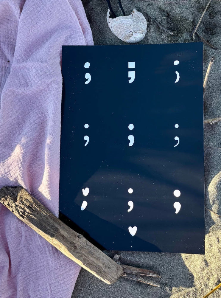 Semicolon print in white on black flatlay displayed on sandy beach, hope collection print by Paper Heart Design NZ