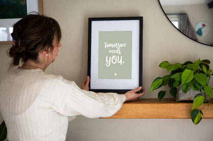 Tomorrow Needs You print in white on sage framed in black with white matt, placed on shelf with plant and mirror behind, raising funds for suicide prevention, designed with love by Paper Heart Design NZ