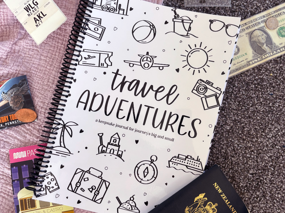 Travel Adventures keepsake journal front cover flat lay on pink and grey fabric with passport, tickets and US money, travel keepsake by Paper Heart Design NZ