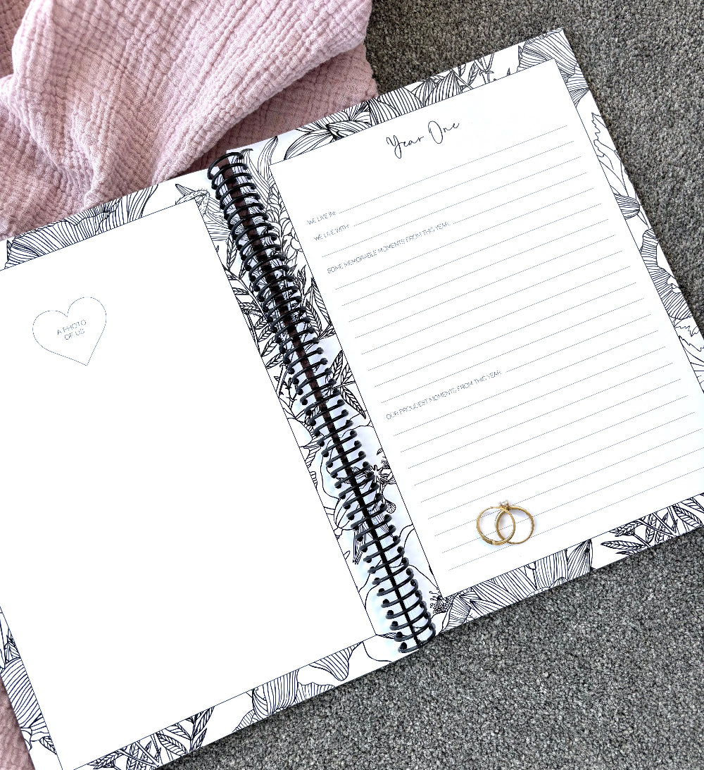 You and Me Anniversary keepsake book open to year one page spread with milestone prompts and photo space, gold rings on pink and grey fabric, by Paper Heart Design NZ