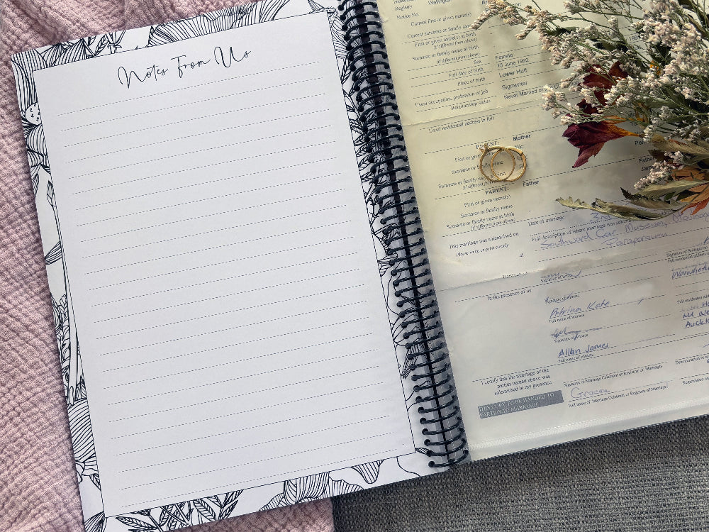 You and Me wedding guest book open to Notes From Us page and back pocket holding marriage license with dried flowers and wedding ring set on pink and grey fabric, by Paper Heart Design NZ