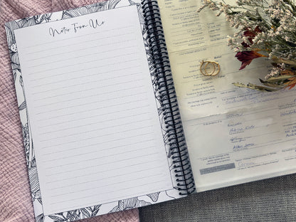 You and Me wedding guest book open to Notes From Us page and back pocket holding marriage license with dried flowers and wedding ring set on pink and grey fabric, by Paper Heart Design NZ