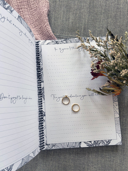 You and Me wedding guest book partially open showing four page prompt spread with gold rings on pink and grey fabric with dried flowers, by Paper Heart Design NZ