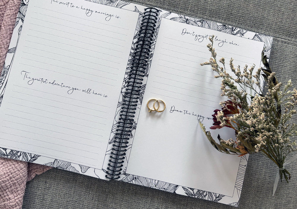 You and Me wedding guest book open to prompt pages with wedding ring set and dried flowers on textured surface, by Paper Heart Design NZ