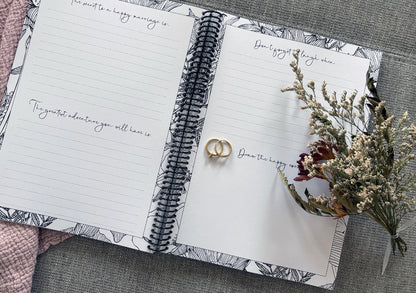 You and Me wedding guest book open to prompt pages with wedding ring set and dried flowers on textured surface, by Paper Heart Design NZ