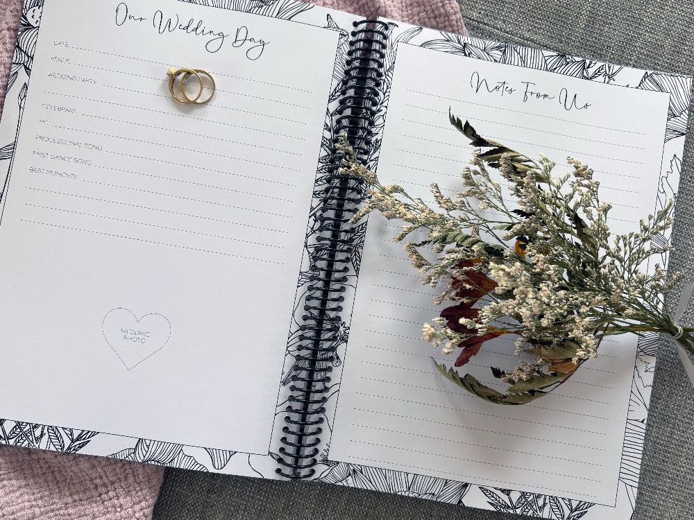 You and Me wedding guest book open to Our Wedding Day and Notes From Us pages with floral design, dried flowers and wedding ring set on textured surface, by Paper Heart Design NZ