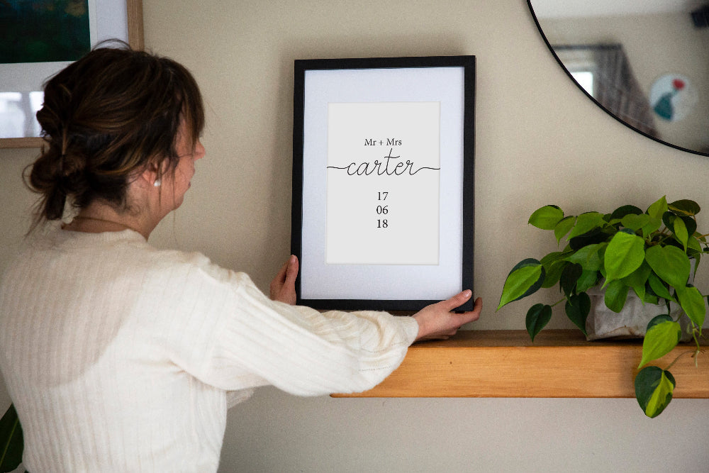 You and Me Wedding Date print in black frame held by brunette woman over shelf with mirror and plant behind, personalised wedding wall art by Paper Heart Design NZ