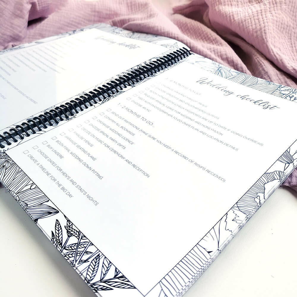 You and Me wedding planner open to wedding checklist pages on pink textured background, NZ wedding planning book by Paper Heart Design